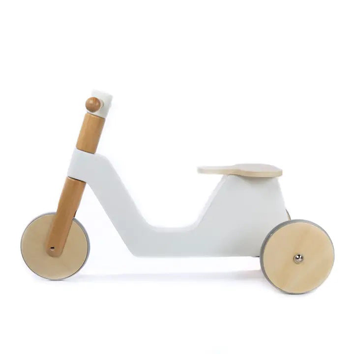 The FLOSS AND ROCK - WOODEN RIDER by GROVE is a minimalist wooden balance bike for toddlers, featuring a white frame, natural wood handlebars, and kid-safe paint. The bike boasts rubber wheels and a low seat with a simple, clean design.