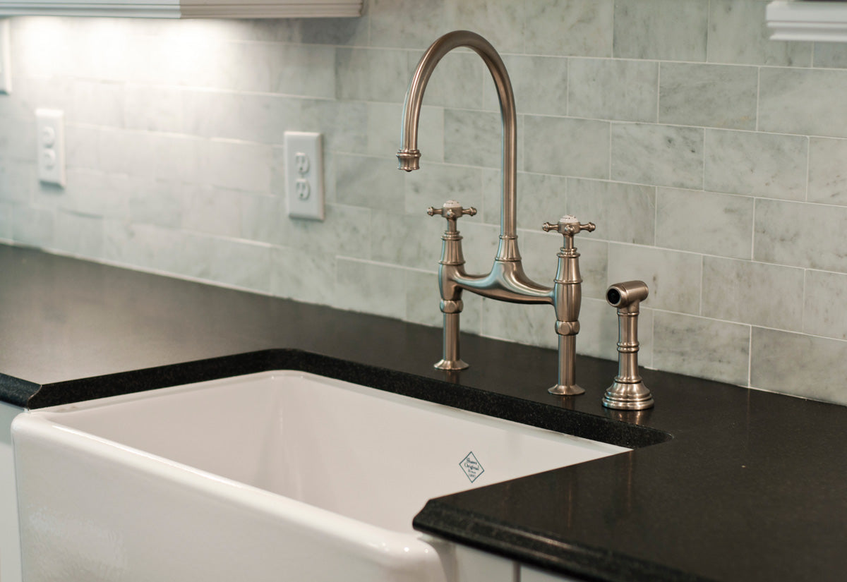 Close-up of a kitchen sink with a white basin, black countertop, brushed nickel faucet, spray nozzle, and a light gray tile backsplash. Electrical outlets are visible on the wall above the counter.