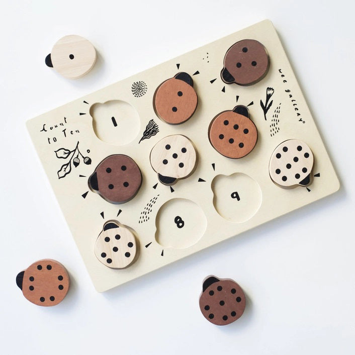 The WEE GALLERY - LADYBUGS COUNT TO 10 TRAY PUZZLE by WEE GALLERY is a wooden puzzle board featuring ladybug-shaped pieces in varying shades from light beige to dark brown. This educational puzzle helps teach counting skills and depicts leaves and twigs alongside the words "Count to Ten" on the left side. Each ladybug is adorned with black dots on their backs to represent number concepts.
