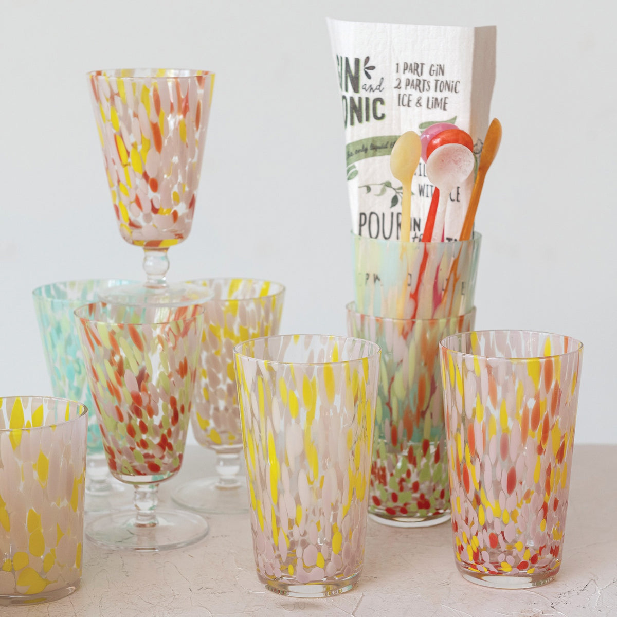 A selection of CREATIVE COOP HAND-BLOWN CONFETTI DRINKING GLASS tumblers, each with red, yellow, and pastel speckles, is displayed on a light surface; a tall glass holds drink stirrers and a cocktail recipe card.