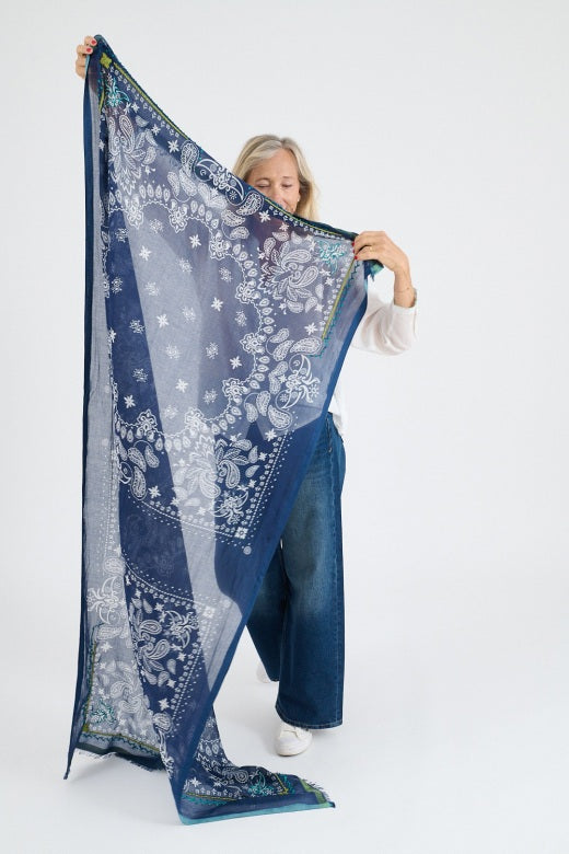 A smiling older woman with blonde hair wears a white blouse and the STORIATIPIC - KIKI SCARF IN MARINE, featuring white and green handmade embroidery. She stands against a plain light background.