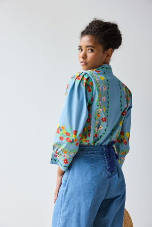 A person wears the HOPE FOR FLOWERS - GIRL SHIRT IN CHAMBRAY FLORAL from HOPE FOR FLOWERS with high-waisted blue jeans, holding a straw hat against a plain, light-colored background.