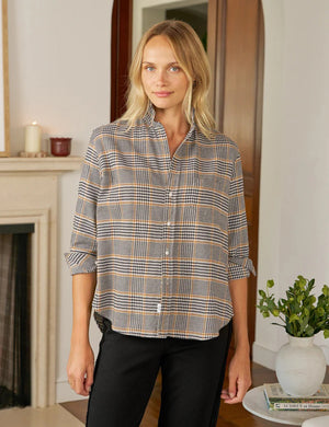A blonde woman stands indoors, smiling softly, wearing the FRANK & EILEEN EILEEN Relaxed Button Up in Italian Flannel Camel Black Plaid and black pants. The cozy room features a fireplace, wooden floors, a plant, and stacked books on a table.