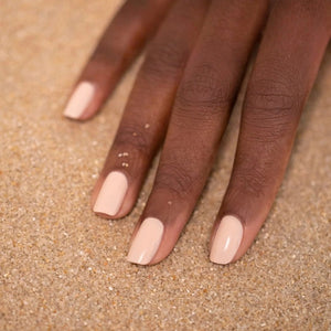 A hand with neatly manicured nails—polished with MANUCURIST - GREEN™ Nail Polish in Pastel Pink by MANUCURIST INC—rests on light brown sand, a few grains clinging to the skin.