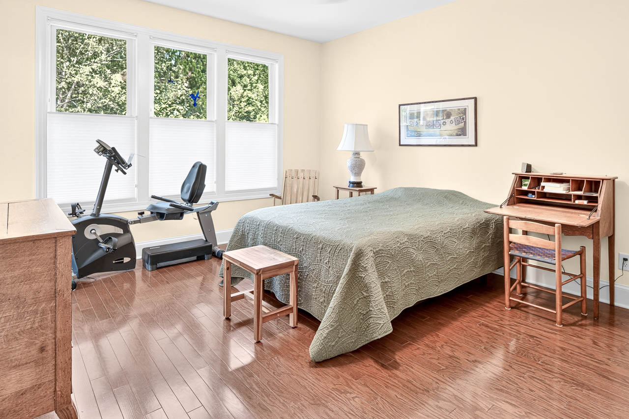 A bedroom with a green bedspread, wooden furniture, a small desk, a lamp, framed art on the wall, and an exercise bike by large windows with white blinds.