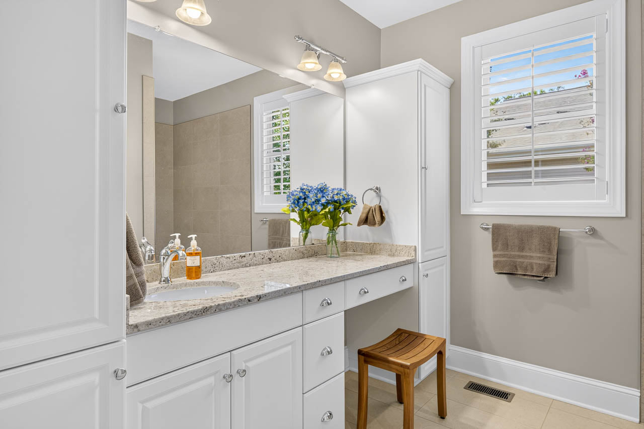 Bright bathroom with white cabinets, granite countertop, mirror, light fixtures, towel racks, wooden stool, and a vase of blue flowers. Window with white shutters is open to outdoor light. Soap bottles are on the counter.