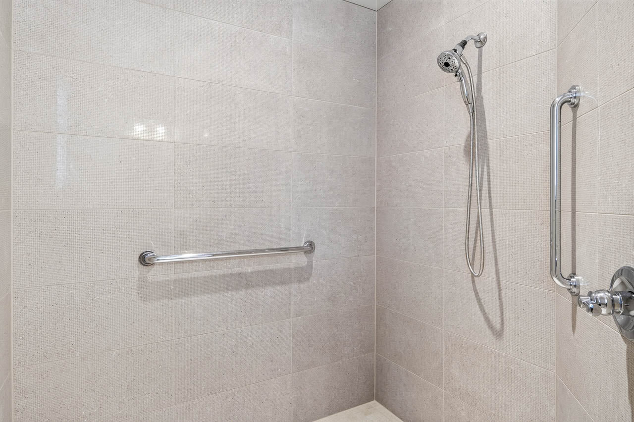 A tiled shower with a handheld showerhead, a horizontal grab bar on the left wall, and a vertical grab bar on the right wall.