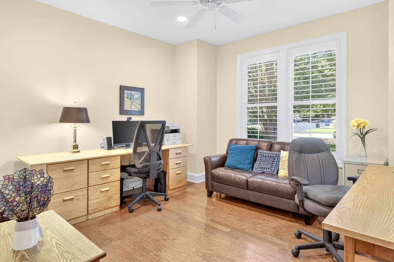 Bright home office with wood floors, two desks, office chairs, a brown leather sofa with pillows, a lamp, flowers, a computer, a printer, and shuttered windows letting in daylight.