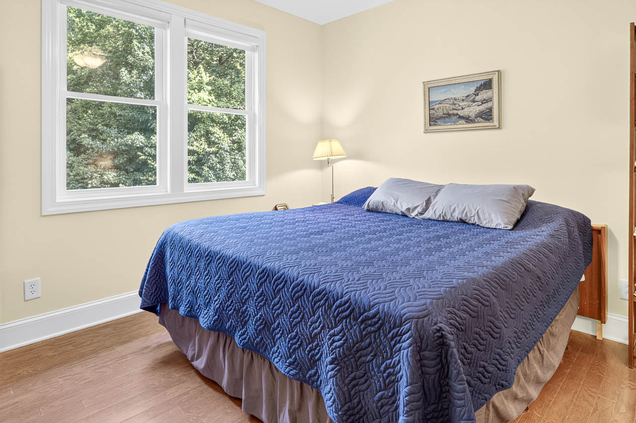 A neatly made bed with a blue quilt and two pillows sits by a window with greenery outside. There is a lamp in the corner and a landscape painting on the beige wall above the bed.