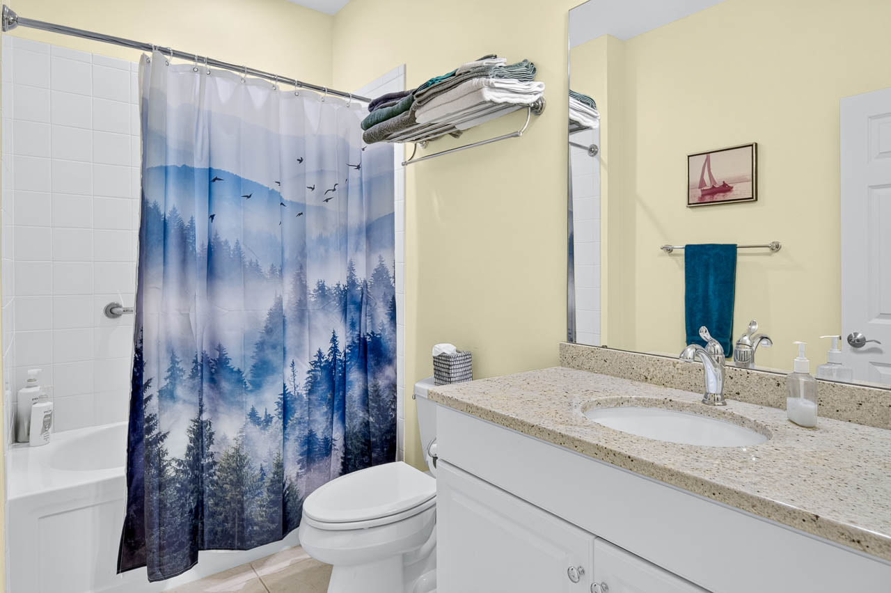 A bathroom with a forest-themed shower curtain, white countertop and cabinets, a wall shelf with folded towels, soap dispensers by the sink, and a small framed sailboat picture on a pale yellow wall.