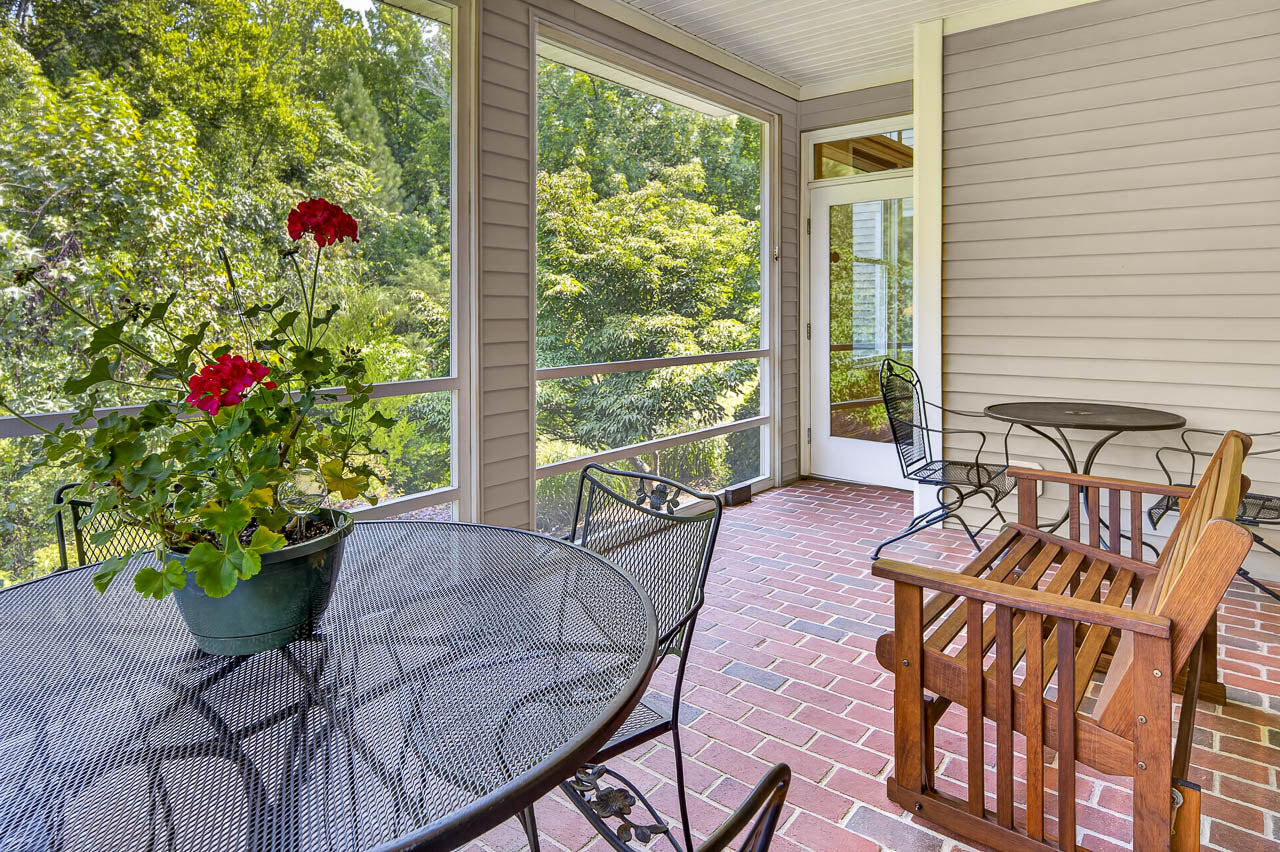 Screened-in porch with brick flooring, black metal table and chairs, wooden bench, small round table, and potted red flowers. Green trees are visible through the screens.