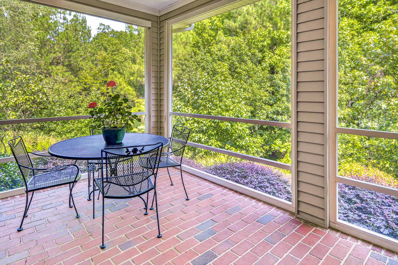 Screened-in porch with a black metal table and chairs, potted flowers on the table, and a view of lush green trees outside. Brick flooring and beige siding complete the bright, peaceful setting.