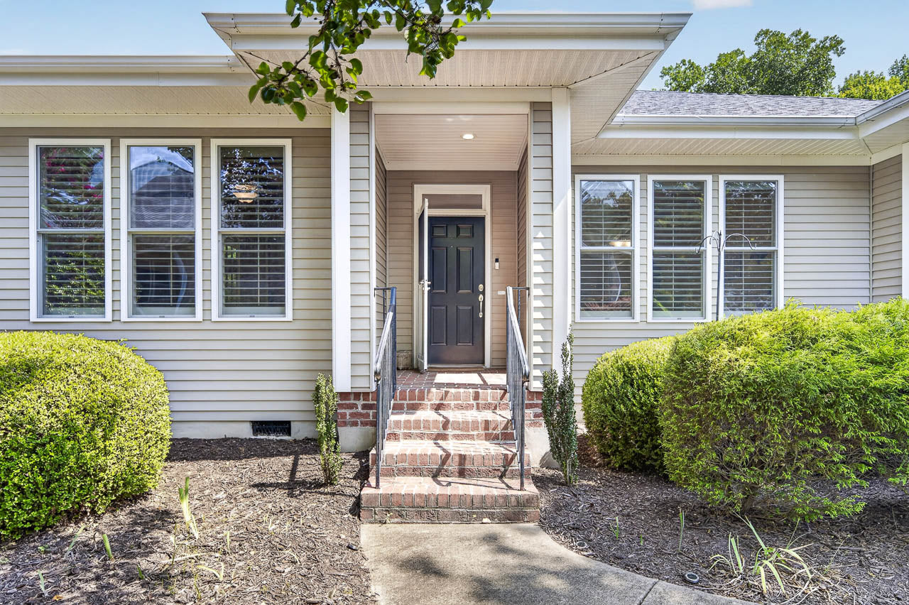 Front entrance of a house with tan siding, white trim, black door, brick steps, metal railings, several windows with shutters, and neatly trimmed bushes in the landscaping.