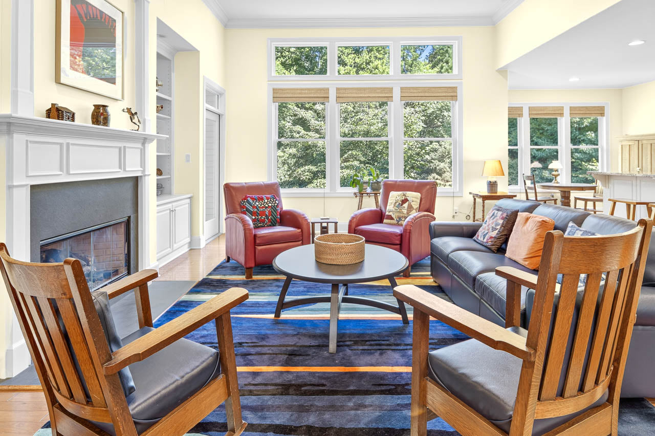 Bright living room with two wooden chairs, a round coffee table, a grey sofa, and two red armchairs. Large windows let in natural light and offer a view of green trees outside. A fireplace and kitchen are visible.