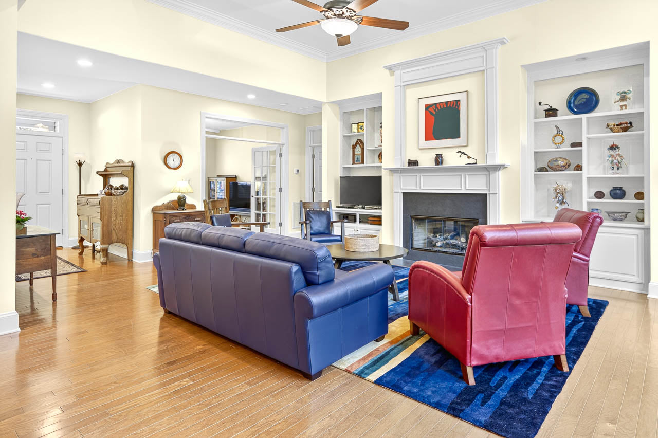 Bright living room with blue and red seating, a fireplace, built-in shelves with decor, wood flooring, and a ceiling fan. A door, desk, and hallway are visible in the background.