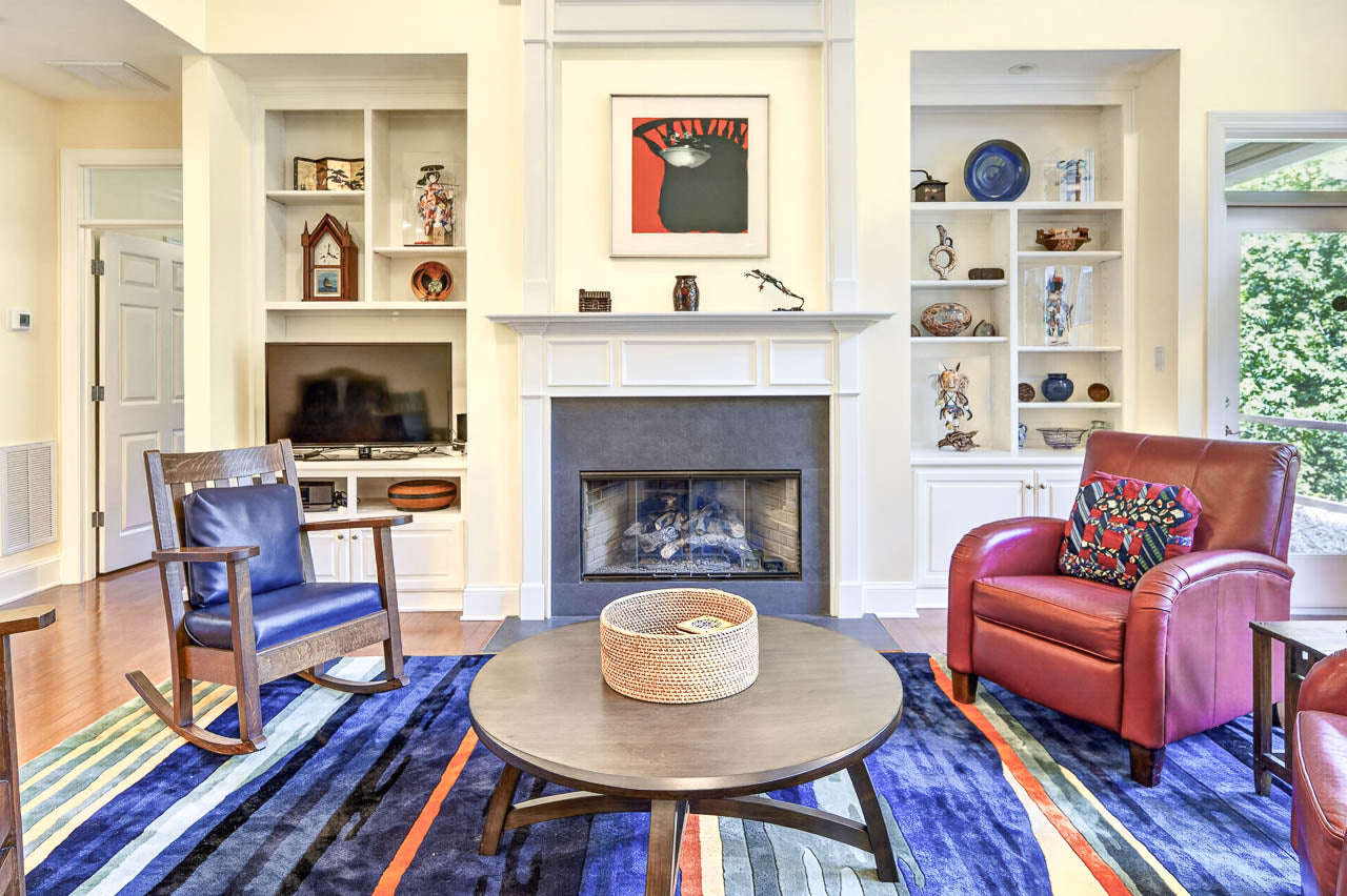 Bright living room with a round coffee table, blue-striped rug, red armchair, blue rocking chair, fireplace, built-in shelves with decor, TV, and large window showing greenery.