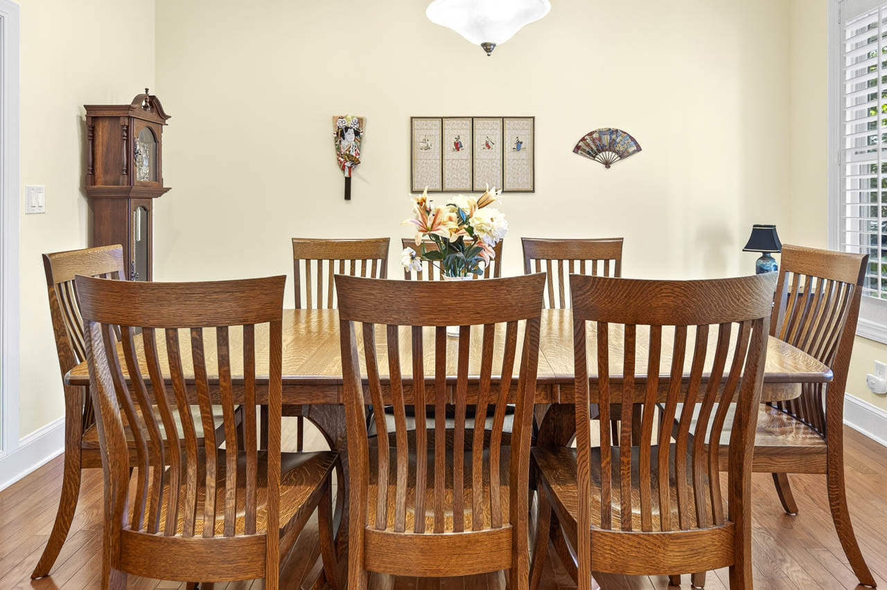 A wooden dining table with eight matching chairs, a flower arrangement centerpiece, and wall decorations in a bright room with hardwood floors and a large window with white blinds.
