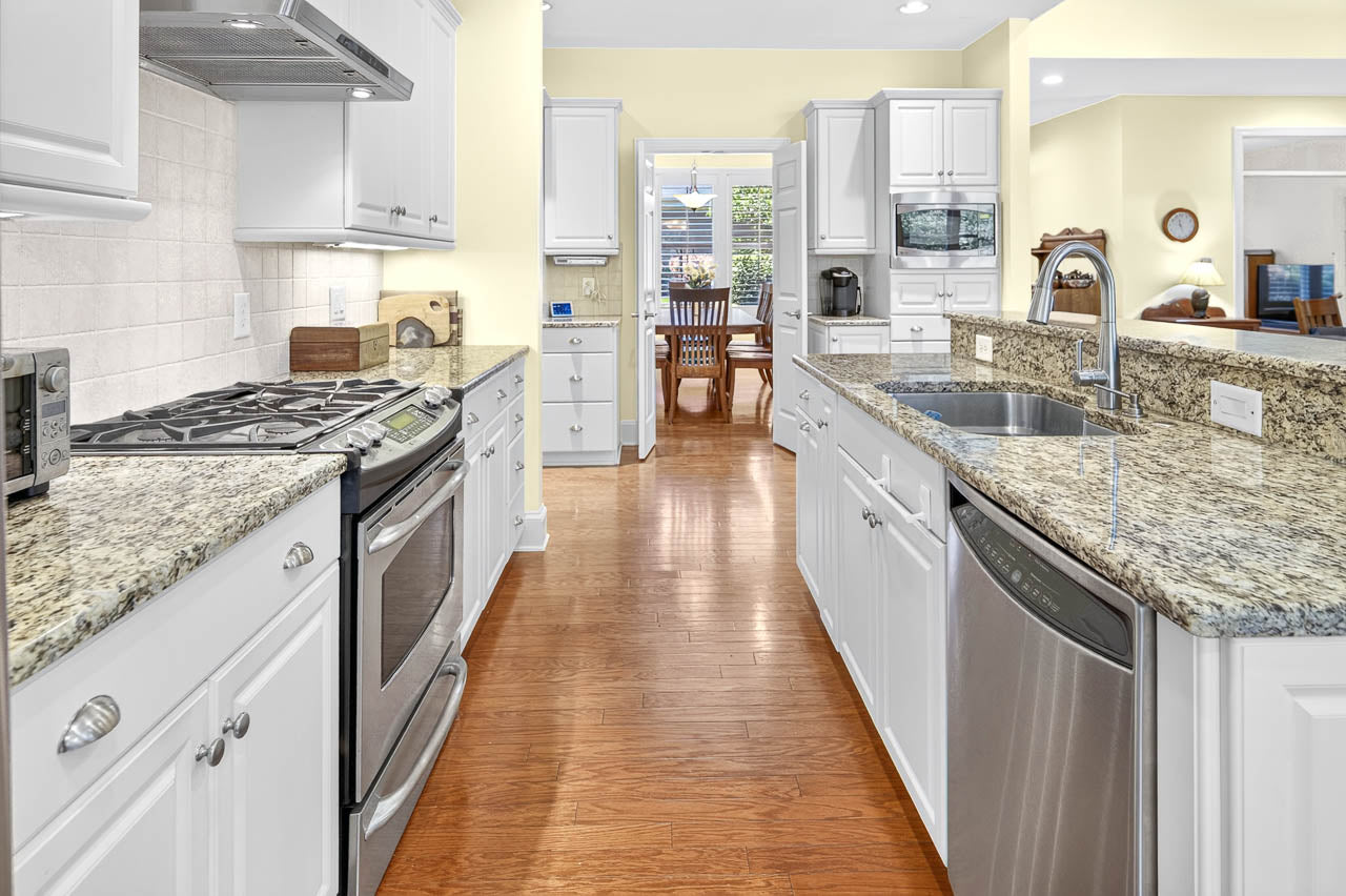 Bright kitchen with white cabinets, granite countertops, stainless steel appliances, and hardwood floors. A dining area with a wooden table and chairs is visible through an open doorway in the background.