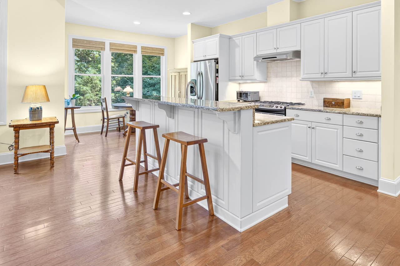 Bright kitchen with white cabinets, granite countertops, stainless steel appliances, and two wooden barstools. Hardwood floor extends to a dining area with large windows, a small table, and a lamp on a side table.