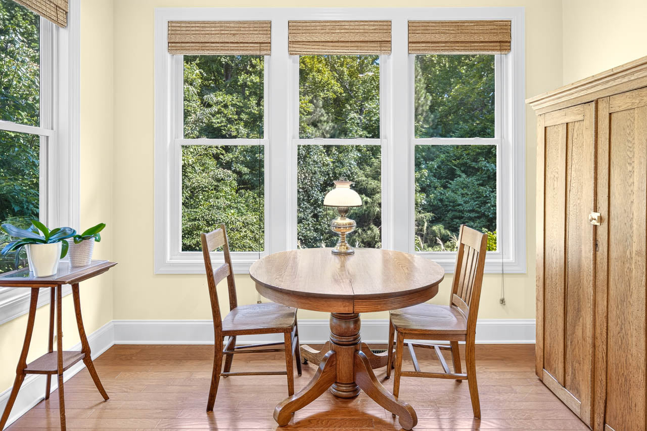 A small dining area with a round wooden table, two chairs, a vintage lamp, and large windows showing green trees outside. A plant sits on a side table next to the windows.