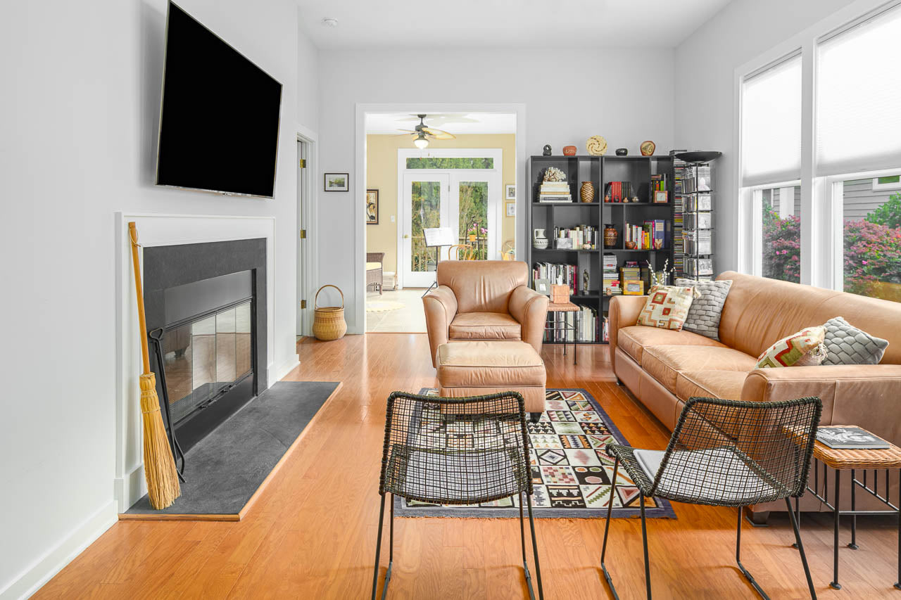 A modern living room with light walls, wood floors, a beige sofa, an armchair, a fireplace, two black chairs, a TV above the mantel, a patterned rug, bookshelves, and large windows letting in natural light.