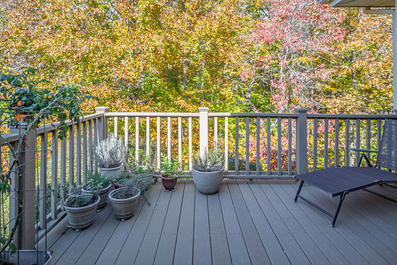 A wooden deck with several potted plants along the railing, a lounge chair to the right, and colorful autumn trees with yellow and red leaves in the background.