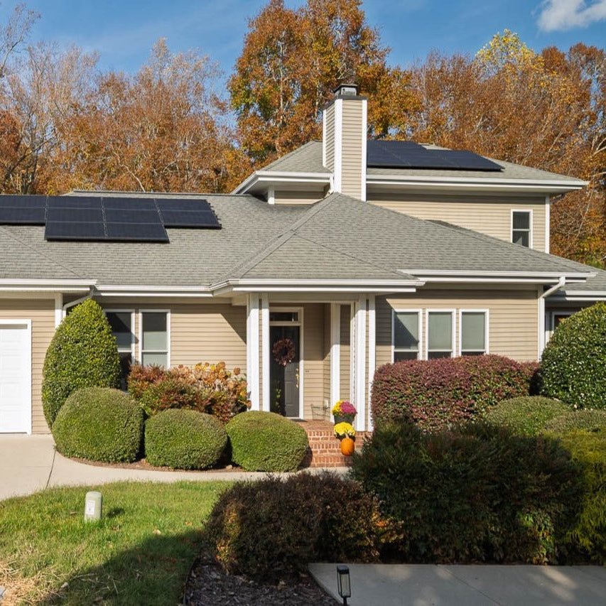 A beige suburban house with two white garage doors, solar panels on the roof, a front porch with potted plants, and neatly trimmed bushes, surrounded by trees with autumn foliage.