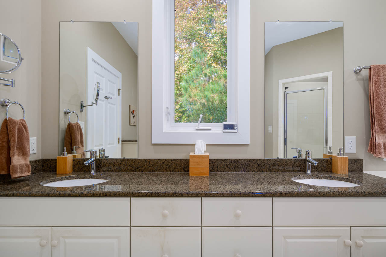 A modern bathroom with a double sink vanity, brown granite countertop, two mirrors, a window between them, beige walls, and neatly hung brown towels. A tissue box sits on the counter between the sinks.