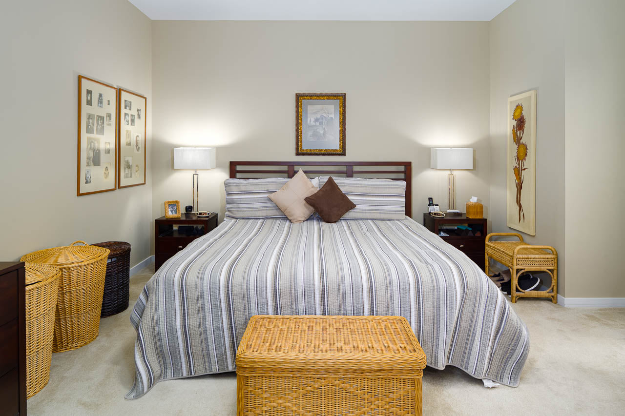 A neatly made bed with striped bedding and two decorative pillows sits between two wooden nightstands with lamps. Wicker baskets and framed pictures decorate the cozy, neutral-colored bedroom.