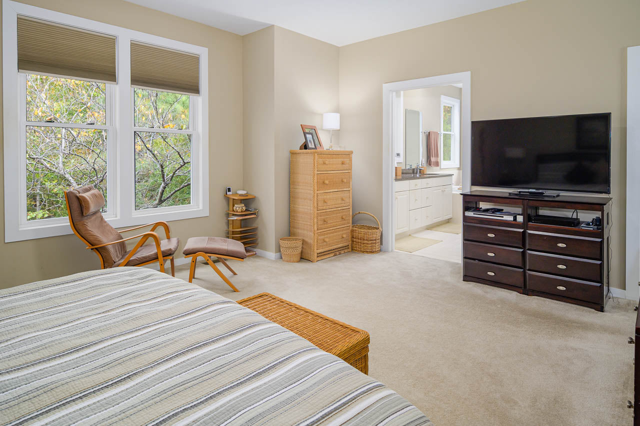 A cozy bedroom with a striped bedspread, wooden furniture, a TV on a dark dresser, and a leather chair by large windows. An open doorway reveals a bright bathroom with a vanity and mirror.