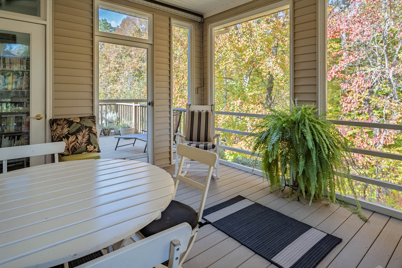 A screened-in porch with a round white table and chairs, a striped armchair, a potted fern, and a view of trees with autumn foliage outside.