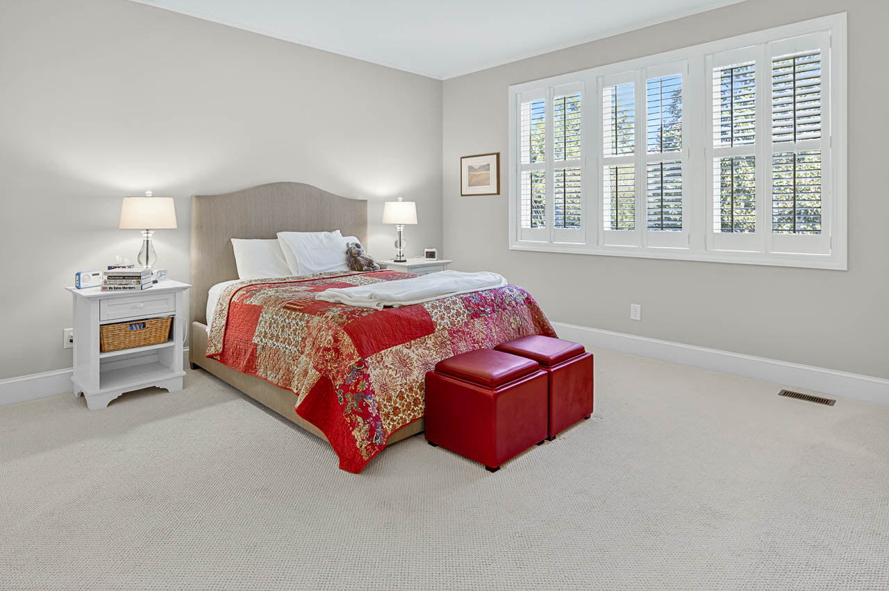 A bright bedroom with a large bed, red patterned quilt, two red ottomans at the foot, white side table with lamp, and a window with white shutters letting in natural light.
