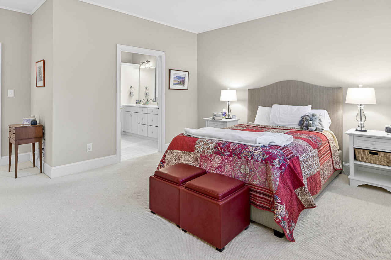 A spacious bedroom with a large bed covered in a colorful patchwork quilt, two red ottomans at the foot, nightstands with lamps on each side, and an open doorway leading to a bright bathroom.