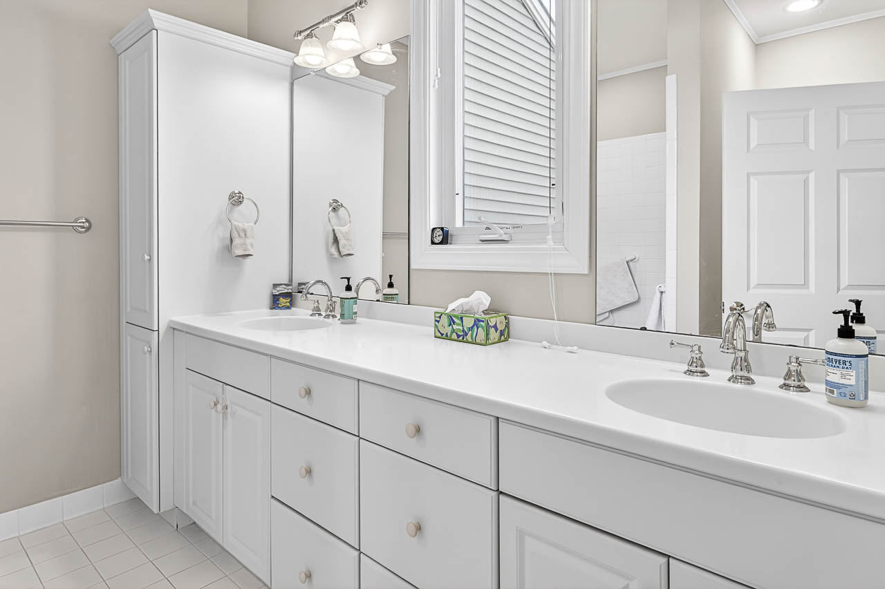 A bright, white bathroom with a double sink vanity, silver faucets, mirrored cabinet, tissue box, soap dispenser, hand towels, and a window with blinds. A glimpse of a shower area is visible through an open door.