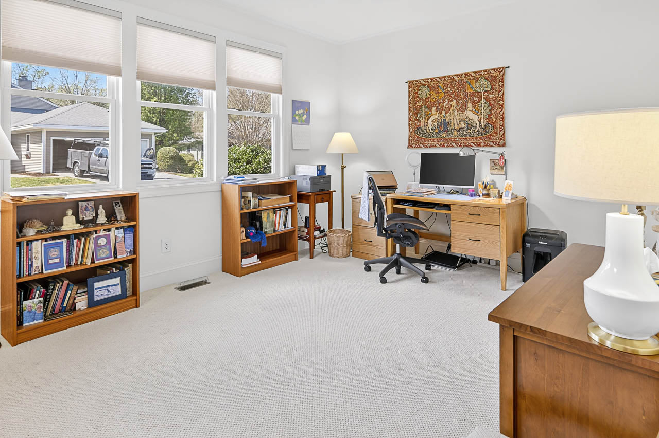 A bright home office with large windows, a wooden desk and chair, a computer, bookshelves filled with books and decor, a floor lamp, and a tapestry hanging above the desk. The room has light carpet and walls.