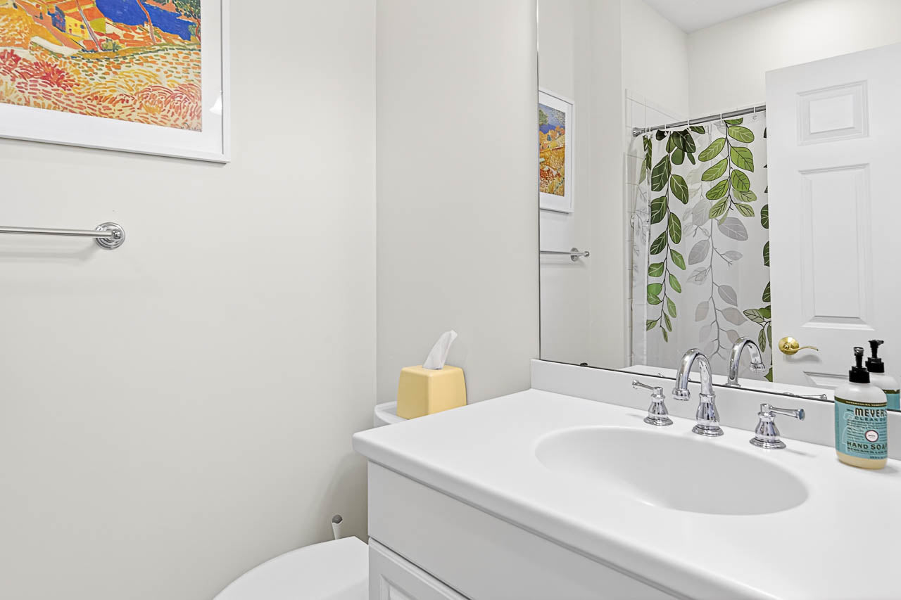 A bright bathroom with a white sink and countertop, a wall mirror, a tissue box, soap dispenser, and a leafy shower curtain. A colorful framed artwork hangs on a pale wall above a towel rack.