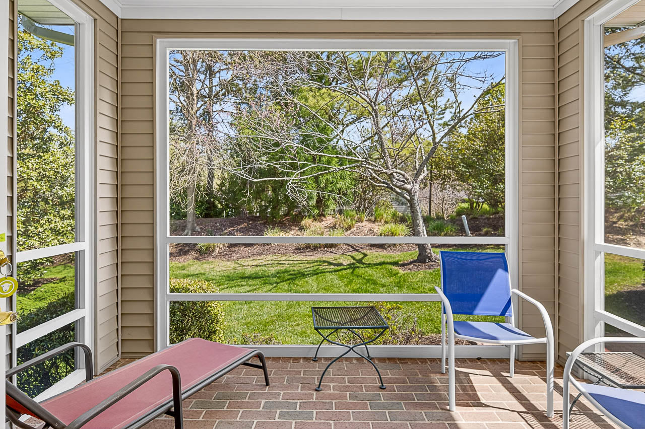 A screened-in porch with brick flooring features a red lounge chair, a blue chair, a white chair, and a small black metal table. The porch overlooks a green, tree-filled backyard on a sunny day.