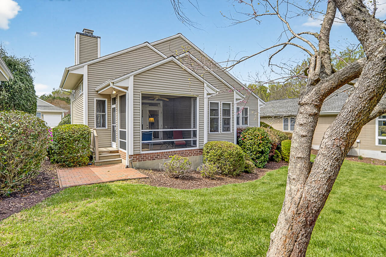 A beige single-story house with a screened porch, surrounded by green bushes and a well-kept lawn, with a tree in the foreground and blue sky above.