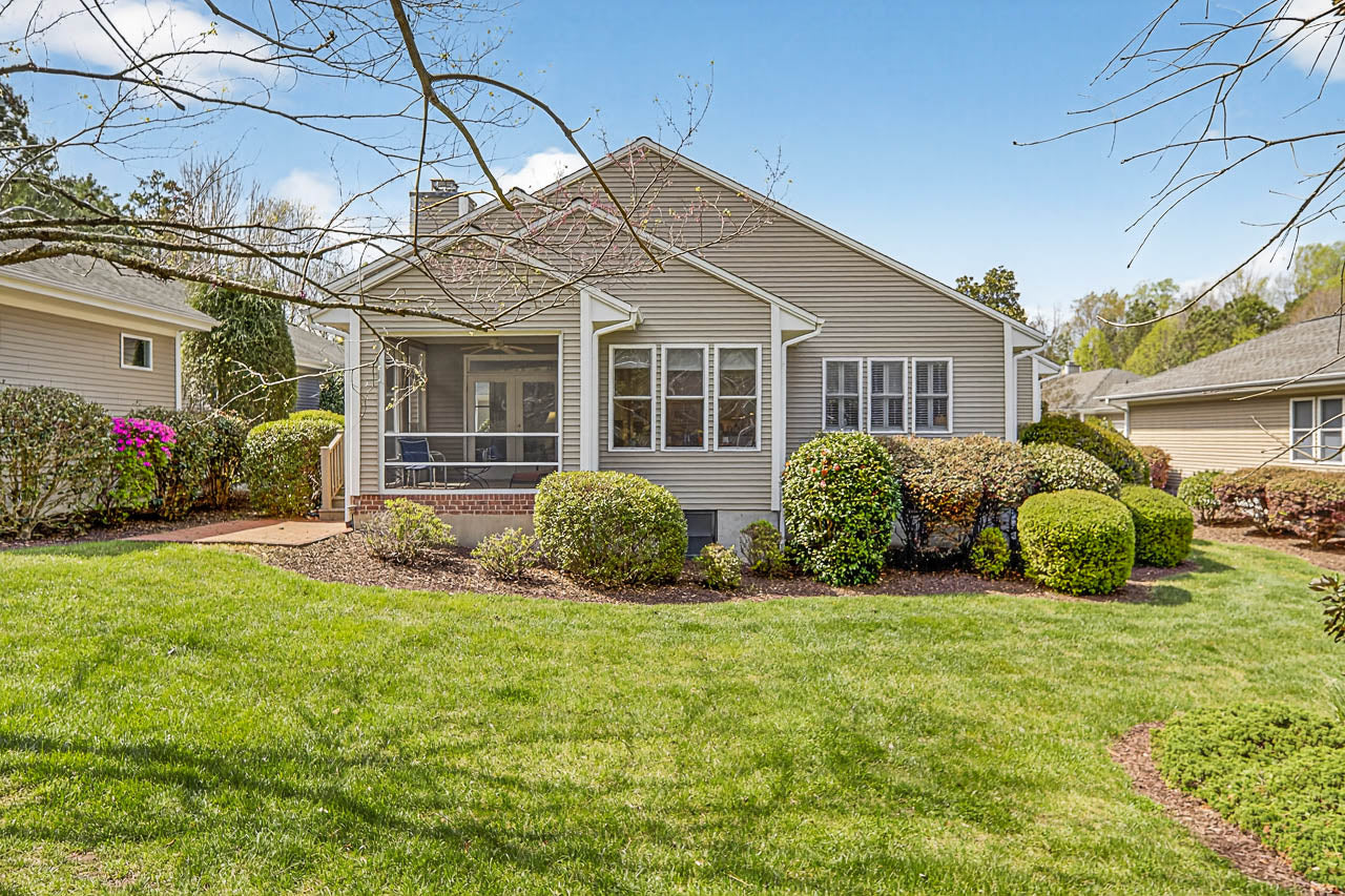 Single-story beige house with white trim, screened porch, and multiple windows, surrounded by neatly trimmed bushes and a green lawn under a blue sky with scattered clouds.
