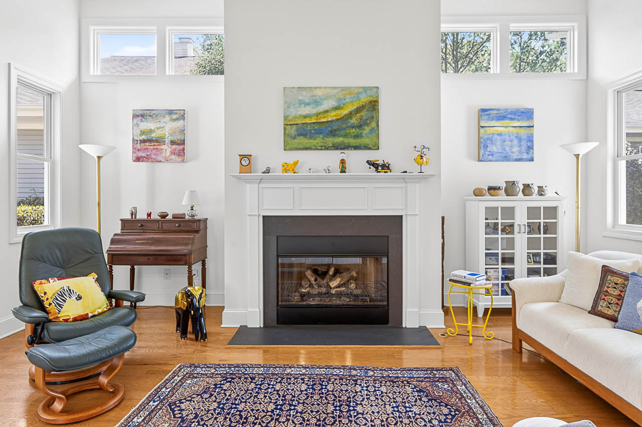 Bright living room with a fireplace, abstract art above the mantel, and tall windows. Room includes a white sofa, a black recliner with a yellow pillow, a wooden desk, and a patterned rug on a wood floor.