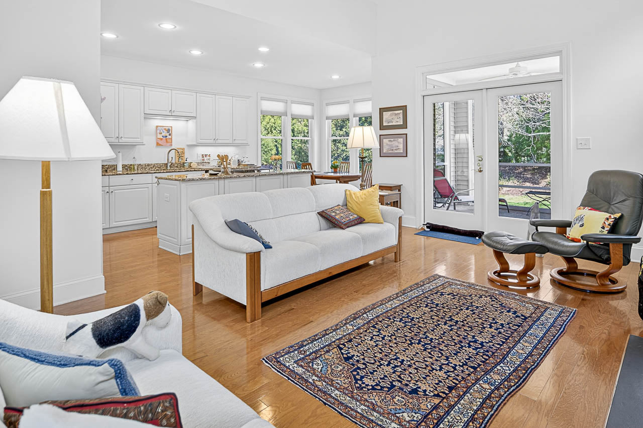 Bright living room with wooden floors, white sofas, a patterned rug, and a lounge chair. The space opens to a kitchen with white cabinets, and large windows show a green, sunlit backyard.