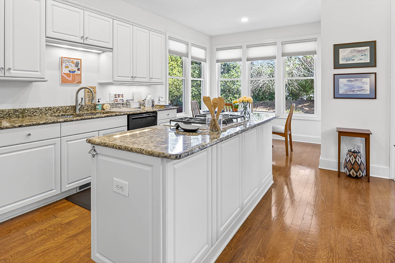 Bright kitchen with white cabinets, granite countertops, and wood floors. A center island holds utensils and plates. Large windows let in natural light, and a dining table is visible near the windows.