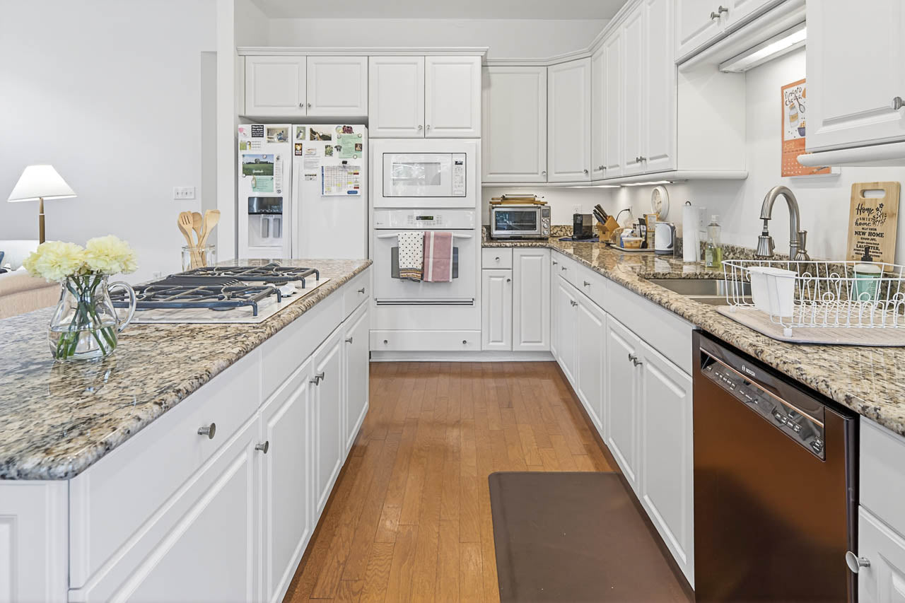 A bright kitchen with white cabinets, granite countertops, stainless steel appliances, a gas stove on an island, and a dishwasher. There's a dish rack by the sink and flowers on the counter. The floor is wooden.