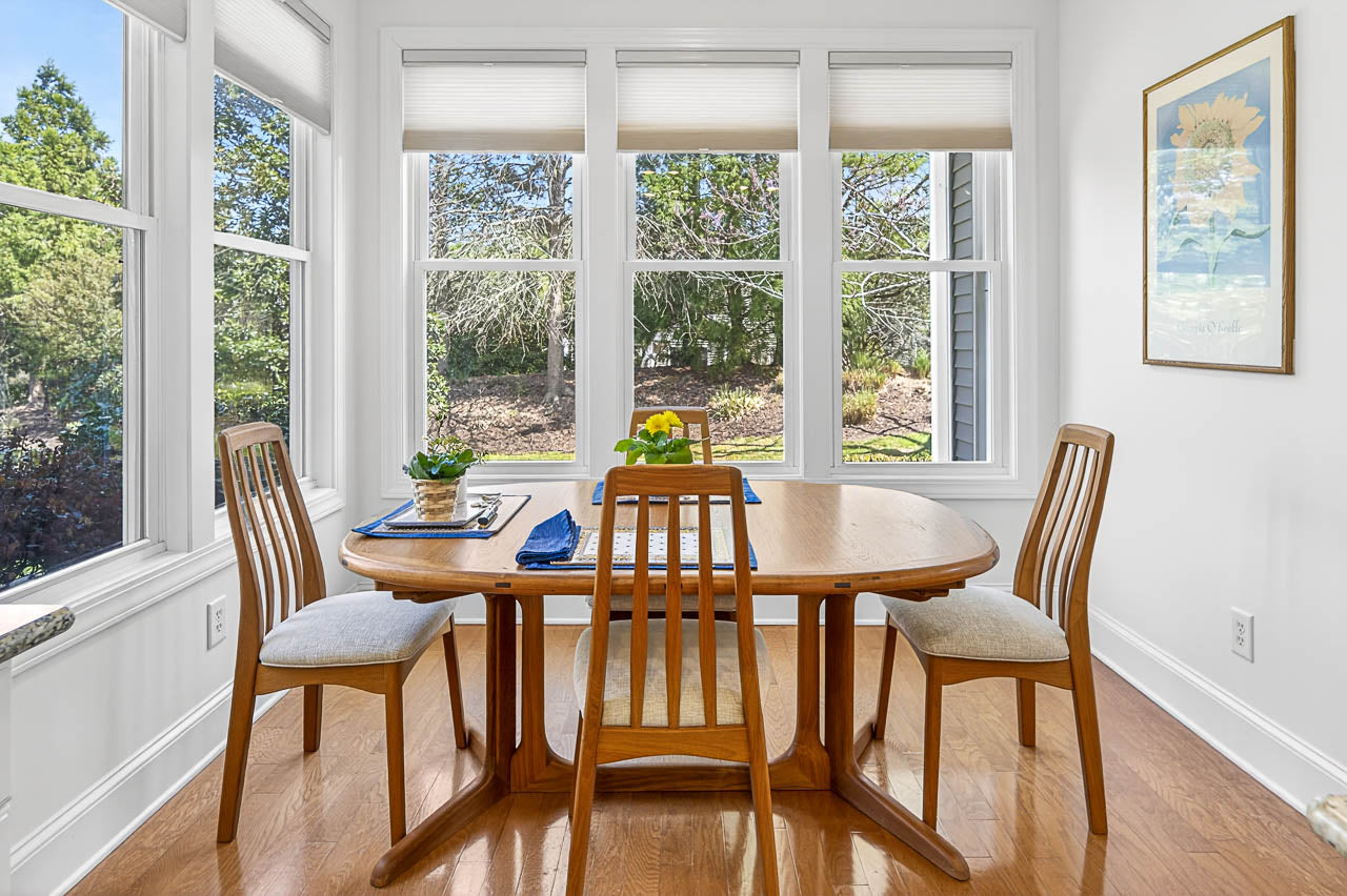 A sunlit dining area with a wooden table and four chairs, set near large windows overlooking a green garden. The table has yellow flowers and placemats, and a sunflower painting hangs on the white wall.