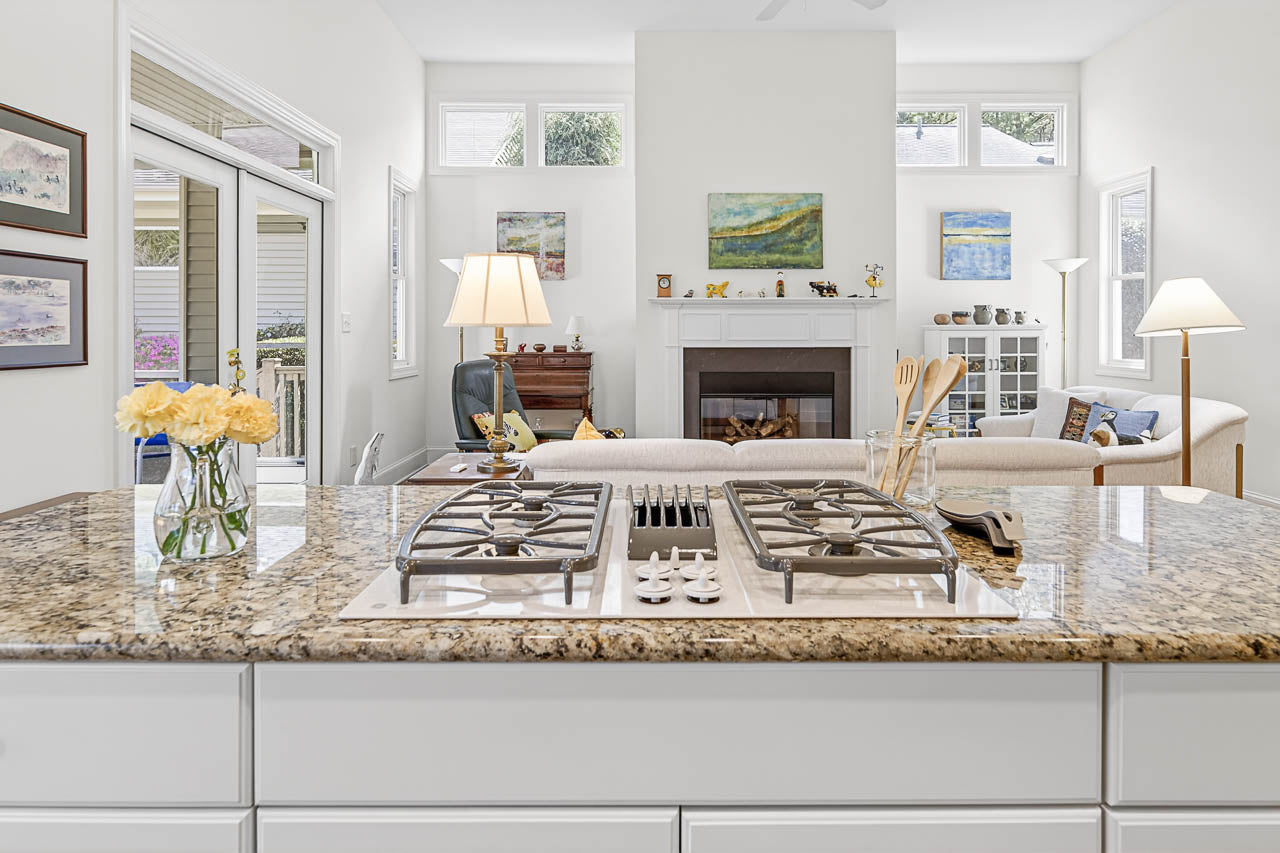 A modern kitchen with a granite island and gas stovetop overlooks a bright living room with a white sofa, fireplace, artwork on the walls, lamps, and large windows letting in natural light.