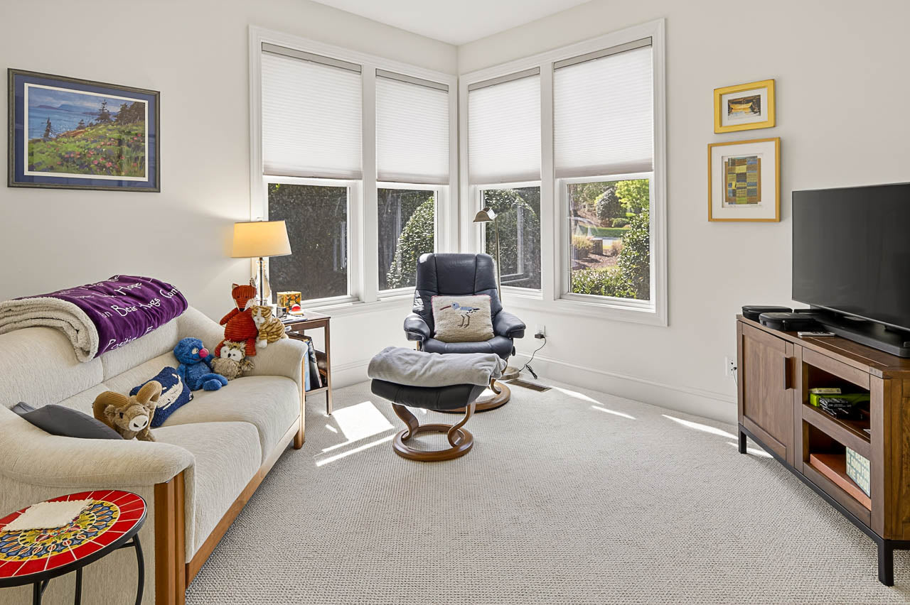 Bright living room with large windows, a white sofa with stuffed animals, a recliner with an ottoman, a TV on a wooden stand, and framed art on the walls. Sunlight fills the cozy space.