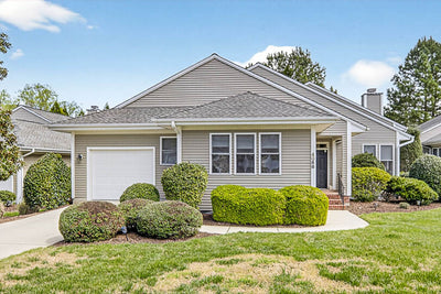 Single-story suburban house with light gray siding, white trim, an attached single-car garage, and well-kept bushes and shrubs in the front yard under a blue sky.
