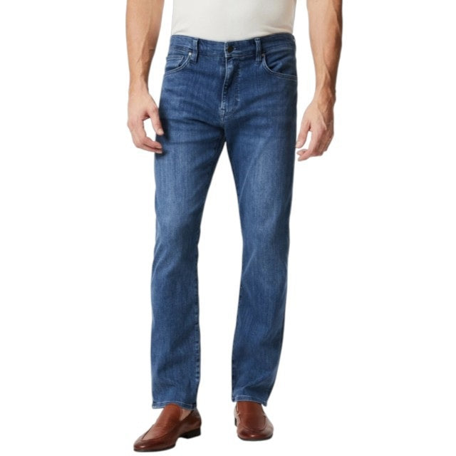 A man wears 34 HERITAGE CHARISMA Relaxed Straight Leg Denim in Mid Urban Wash, a white shirt, and brown leather shoes, standing against a plain white background.