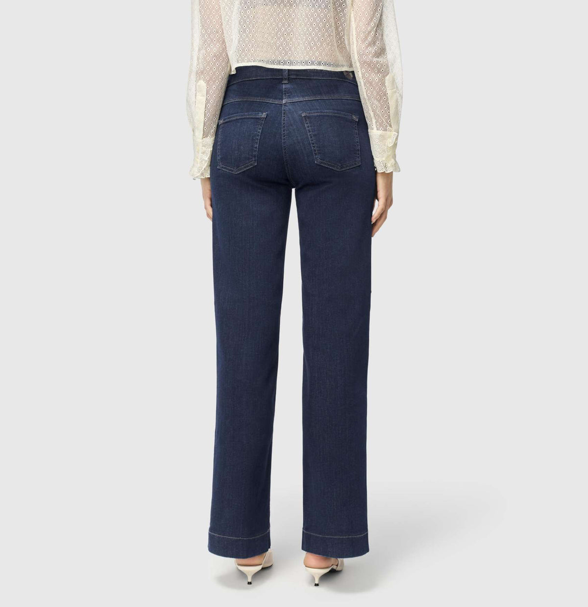 A person with light brown hair stands against a plain background, wearing a sheer, ruffled, long-sleeved white blouse and MAC JEANS - DREAM WIDE AUTHENTIC by MAC in dark blue. They&#39;re paired with comfortable white shoes.
