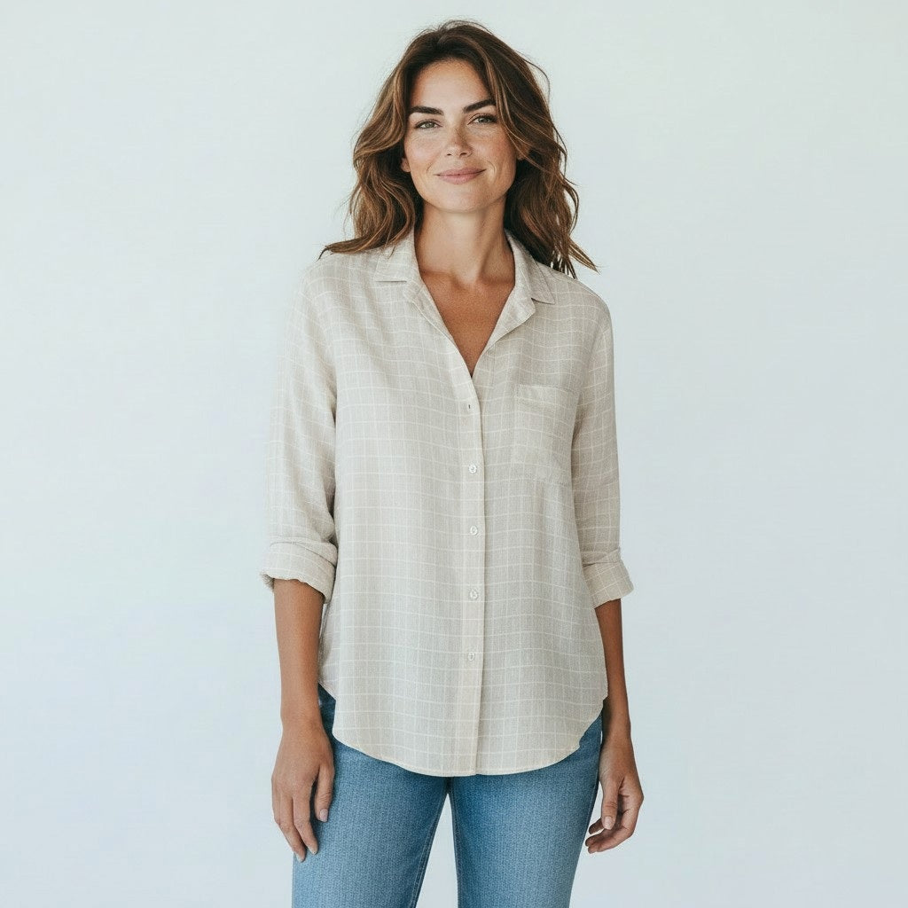 A woman with brown hair, smiling on a sunlit cobblestone street with blurred outdoor café tables in the background, wears the FRANK & EILEEN EILEEN Relaxed Button Up in Italian Cotton Tan with Textured Grid paired with jeans.